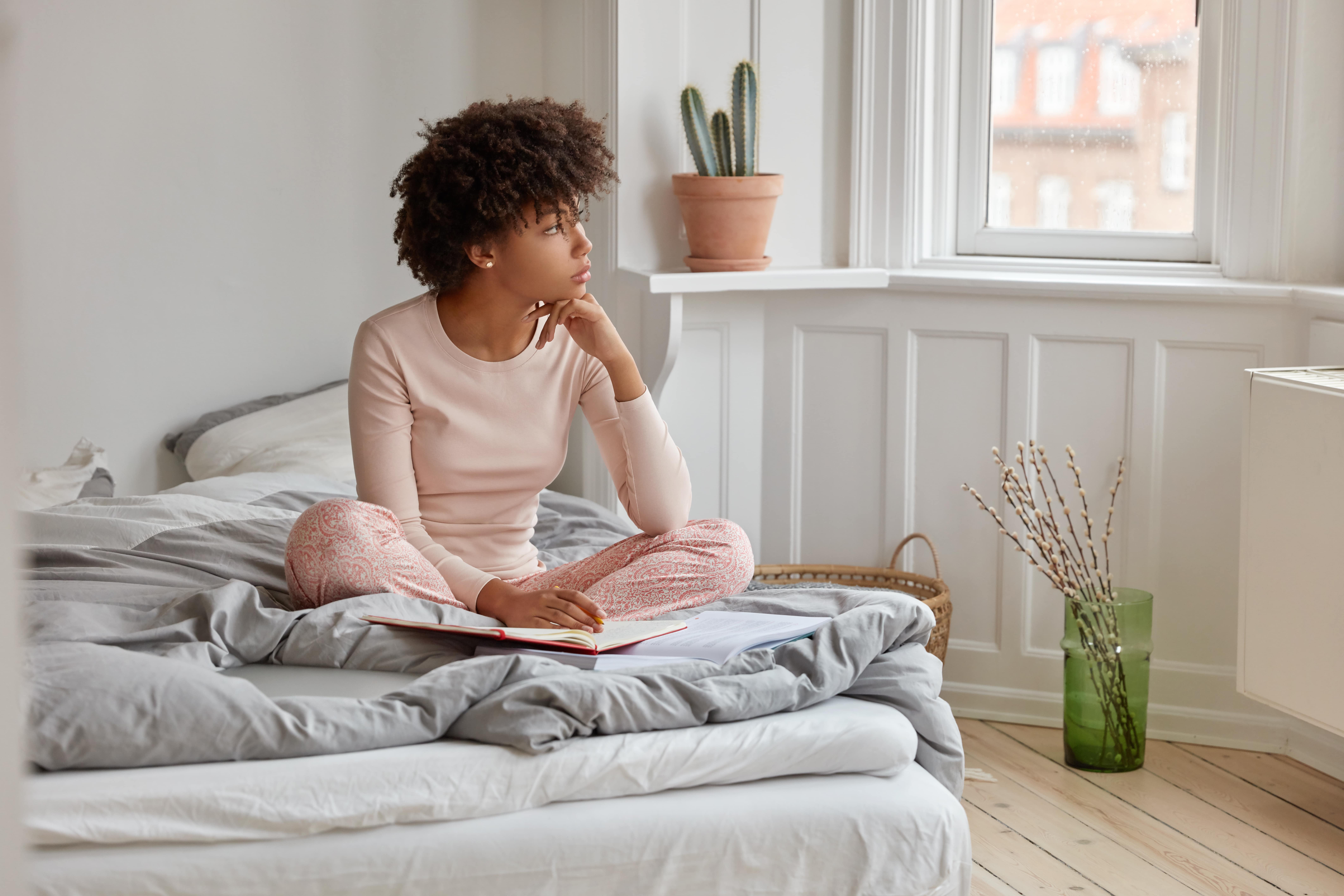 A lady sitting cross-legged on her bed in the morning, looking out of the window while thinking about her sleep last night as she fills out her sleep diary