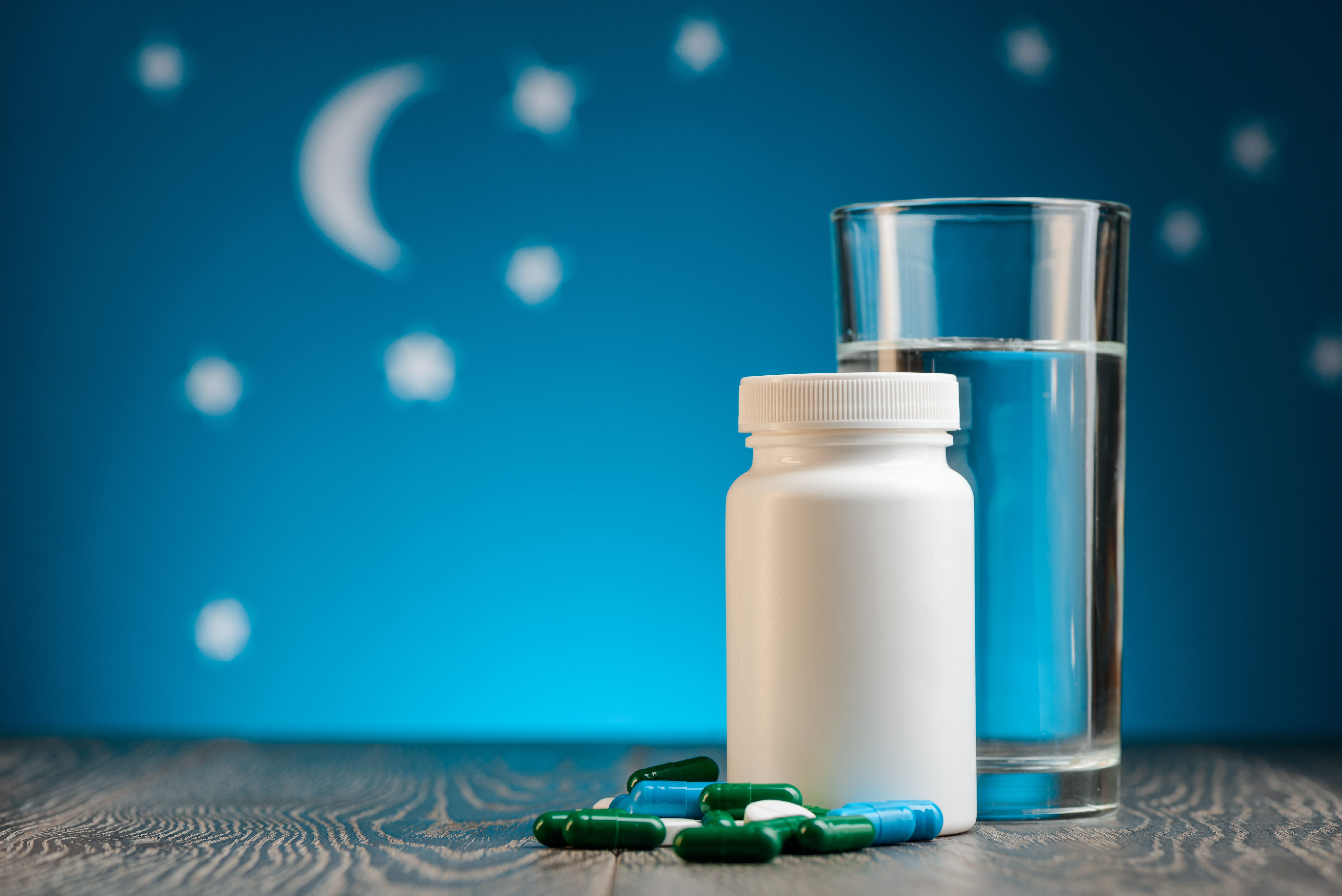 A white tablet container with a scattering of sleep supplements and a glass of water positioned next to it. The background is a child-like night time image featuring stars and a crescent moon against a deep teal-blue background.