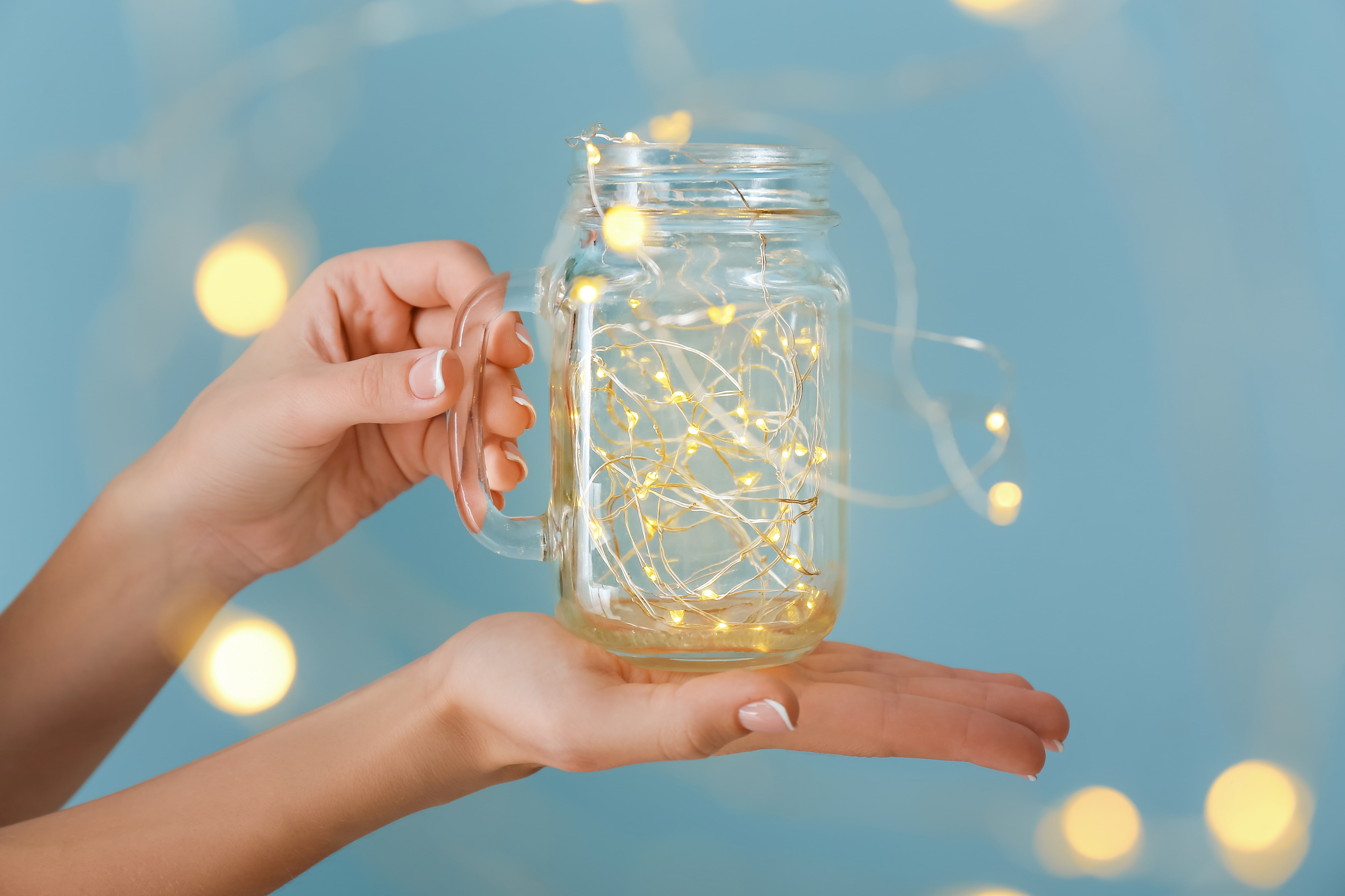 A lady holding a mason jar filled with a warm yellow string of fairy lights against a blue wall.