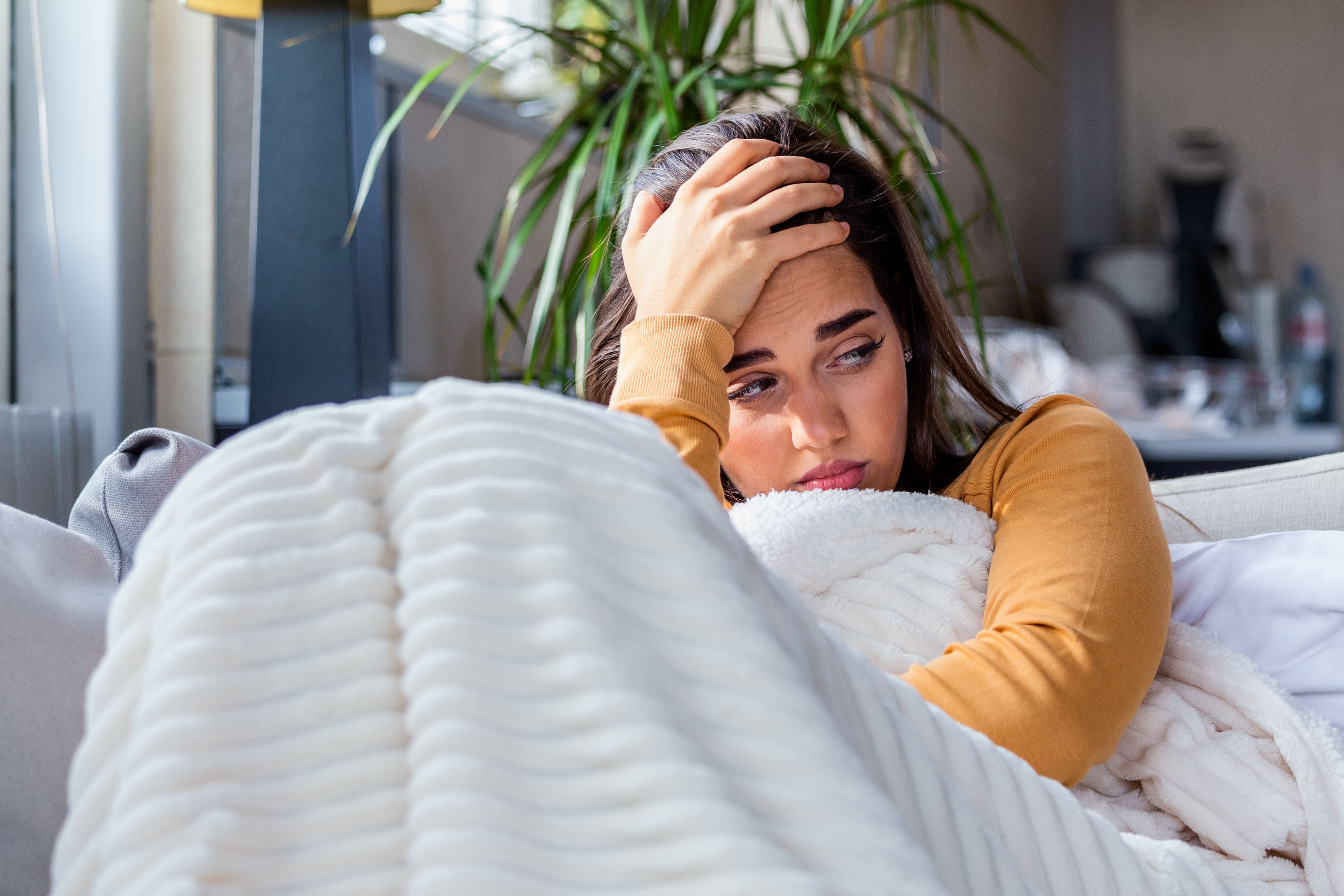 A photo of a lady sat on a sofa with her head in her hands after a bad night's sleep. She is wrapped in a blanket and has an air of distress about her.