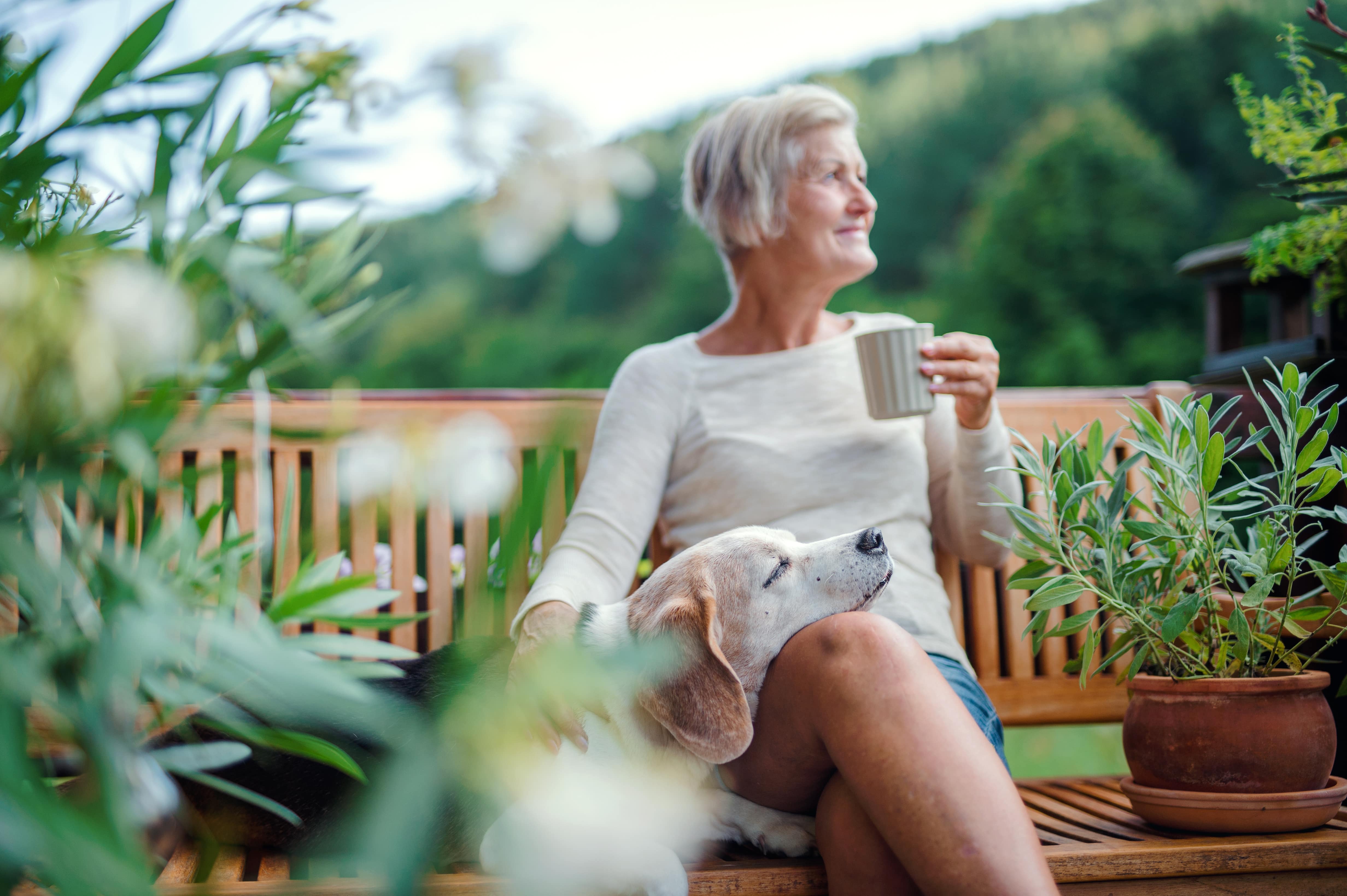 A lady sat on a bench in her garden enjoying a morning cup of coffee while her dog snoozes beside her resting its head on her lap.