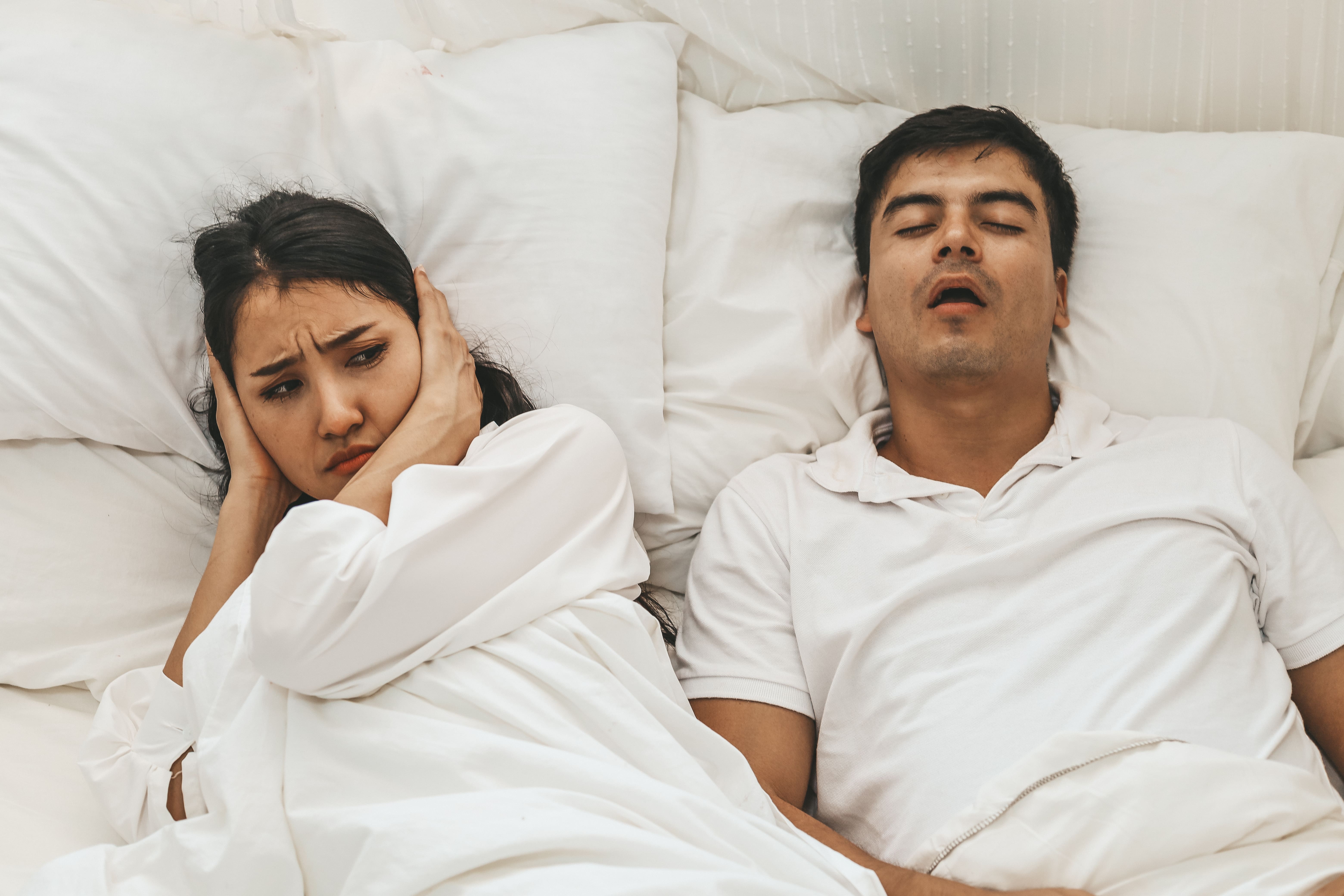 A woman covering her ears while her partner snores as he sleeps next to her.