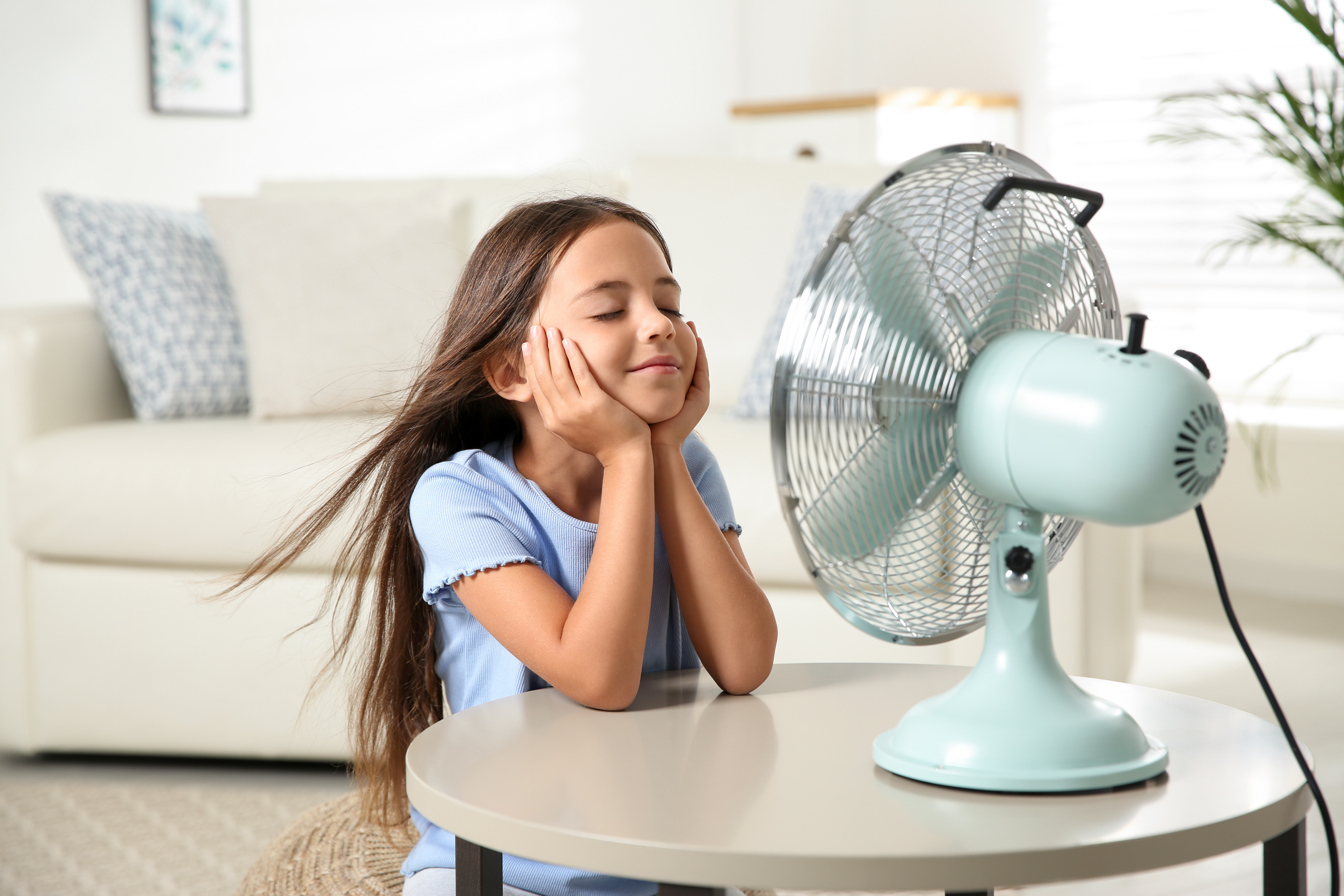 A little girl standing in front of a desk fan, resting her chin in her hands as she cools off on a hot summer's day