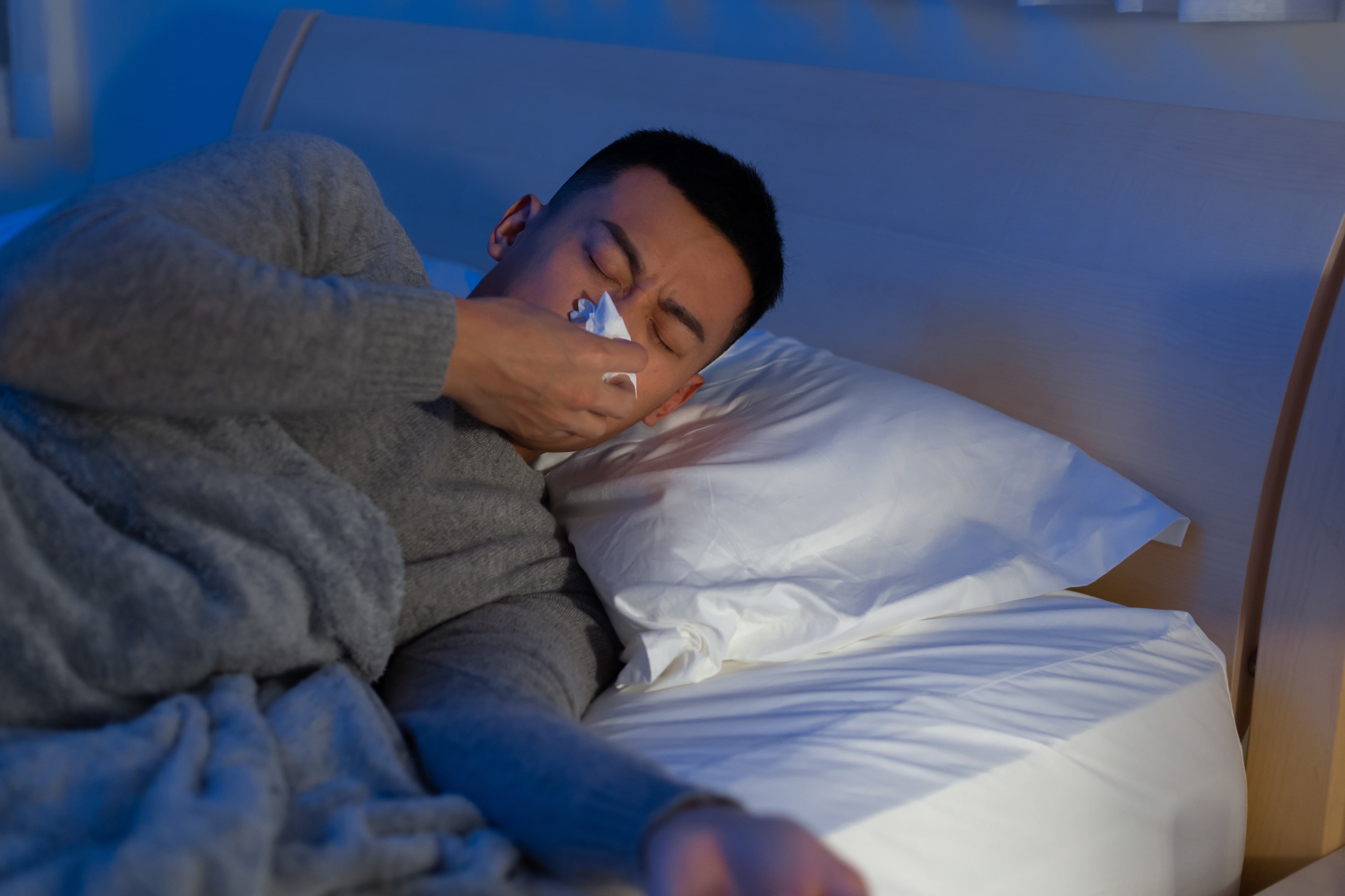 A man blowing his nose while lying in bed and trying to get to sleep while suffering from hay fever