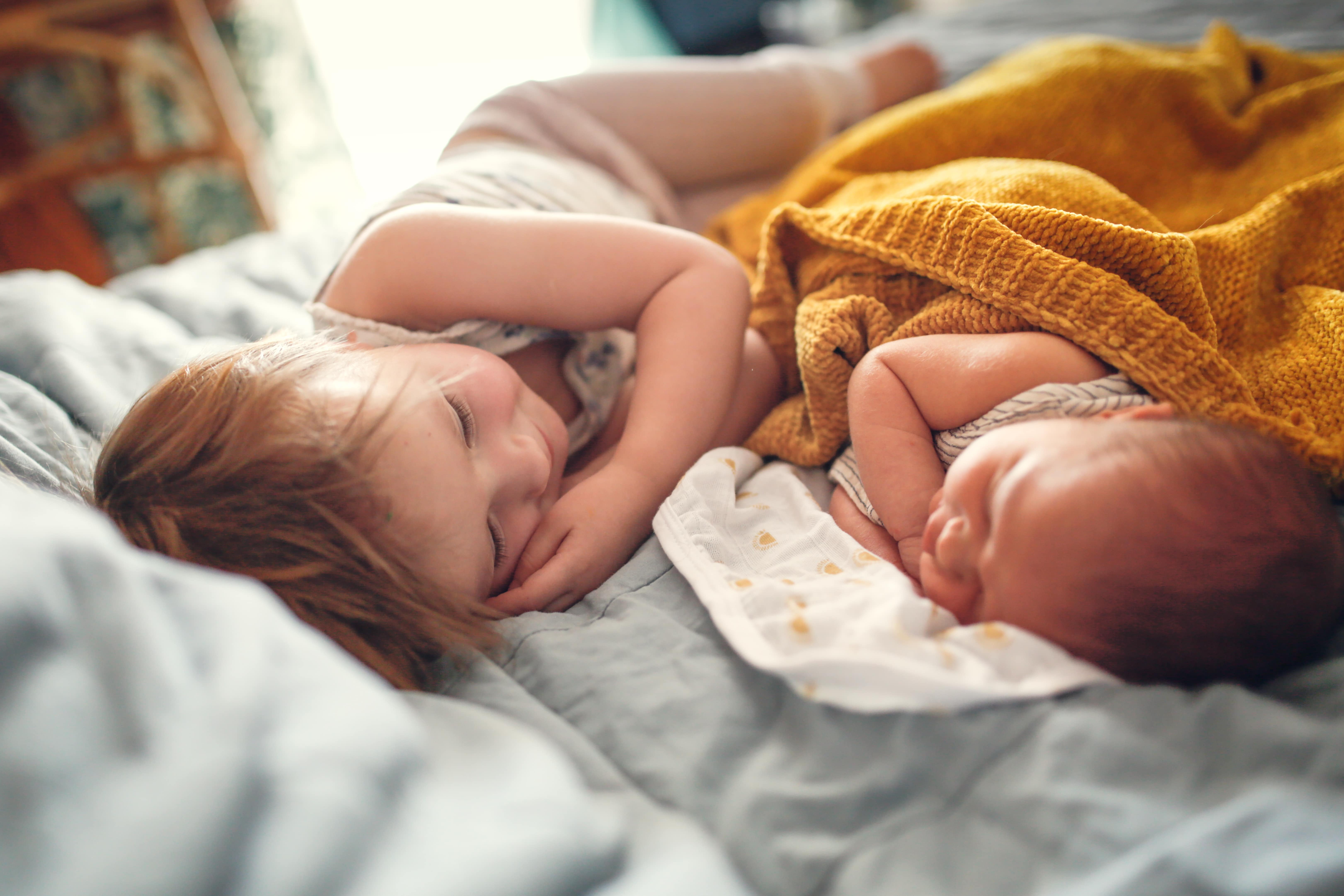 A little girl and her baby brother lying side by side on a comfy duvet. The baby is sleeping while the big sister looks at him lovingly.