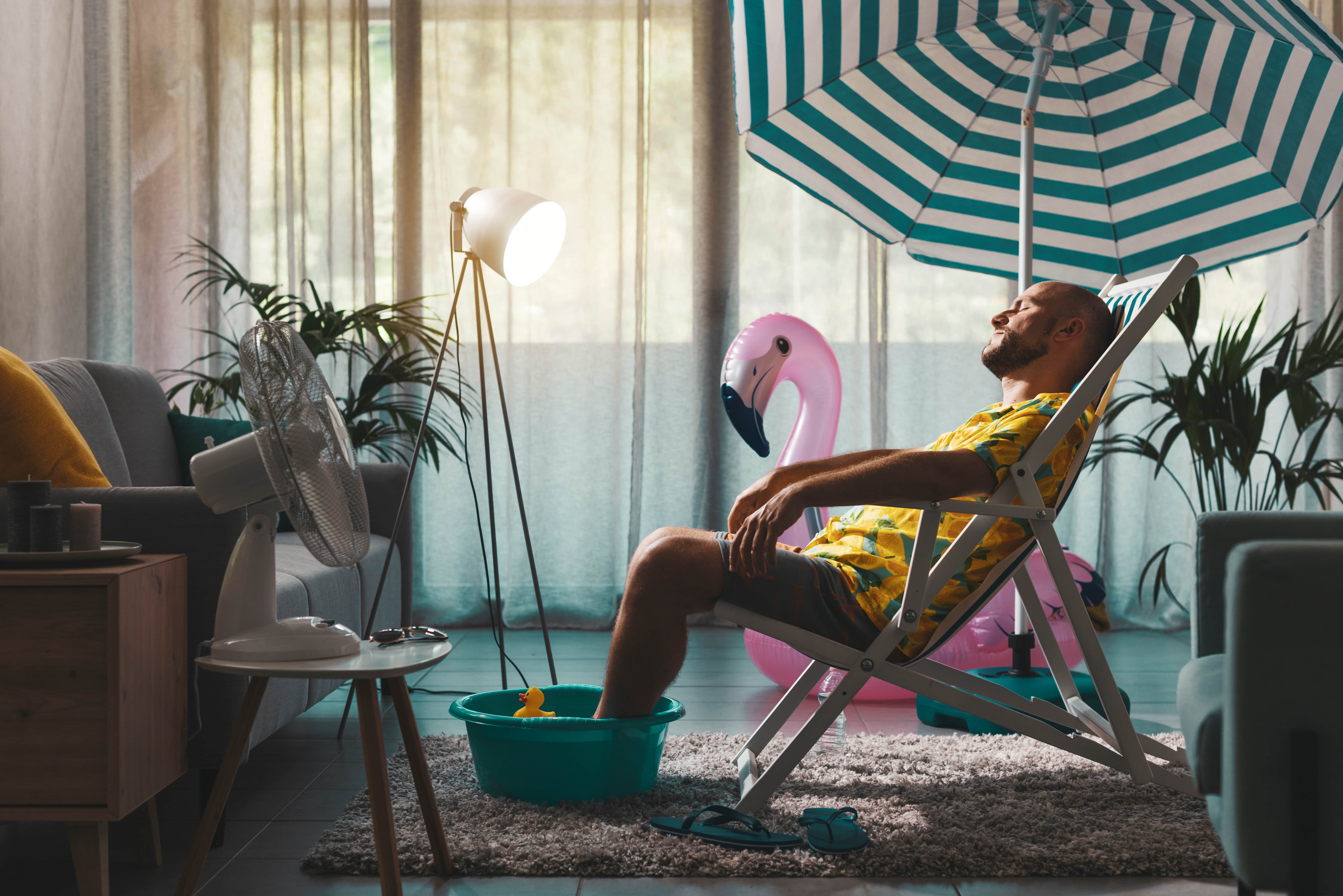 A man sitting on a sun lounger below a parasol indoors. Beside him is an inflatable pink flamingo and a strategically placed fan. He also has his feet in a washing up bowl filled with cold water