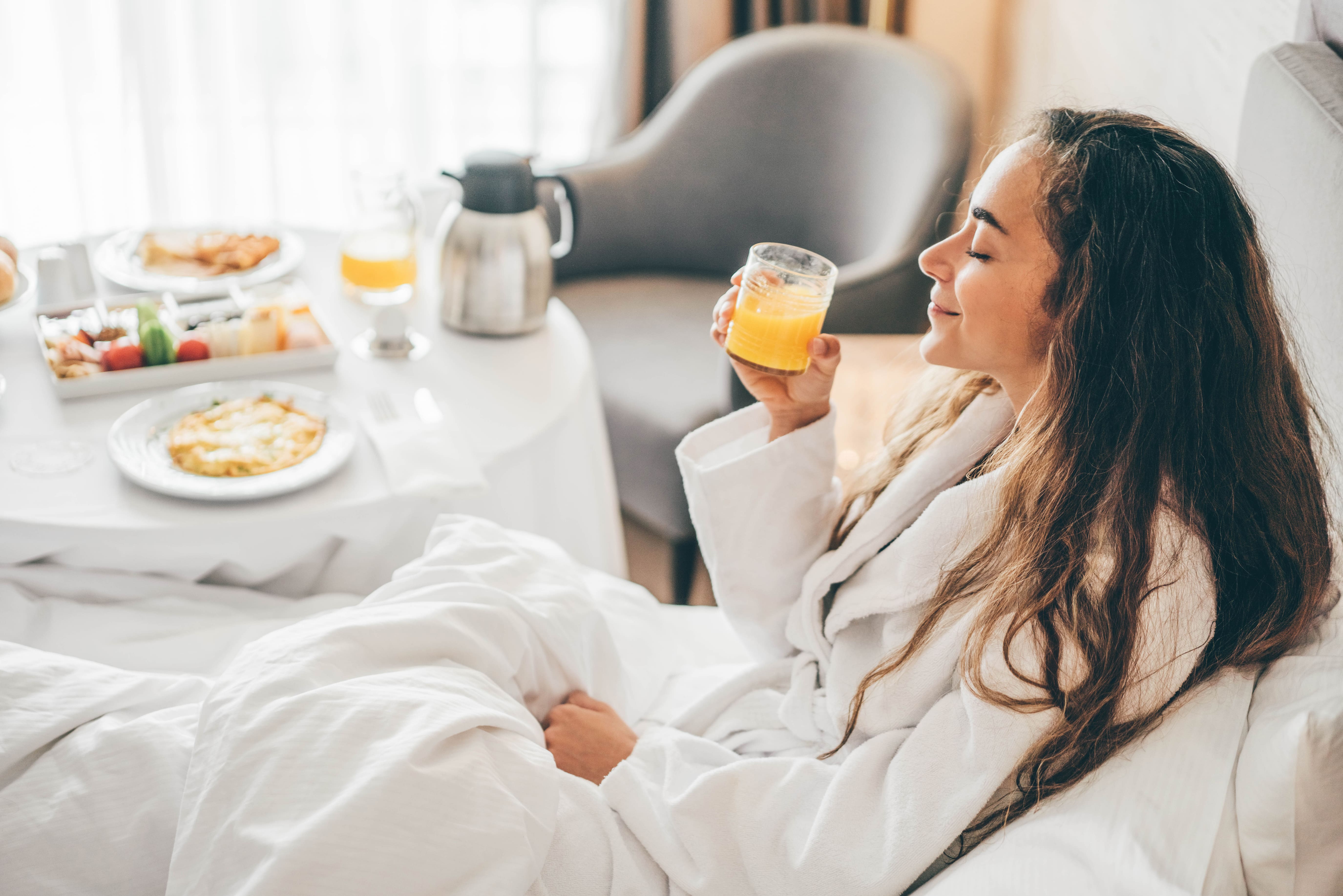 A woman sat in bed with her eyes closed, a glass of orange juice in her hand, and a smile on her face. In the background is a table filled with breakfast delights.