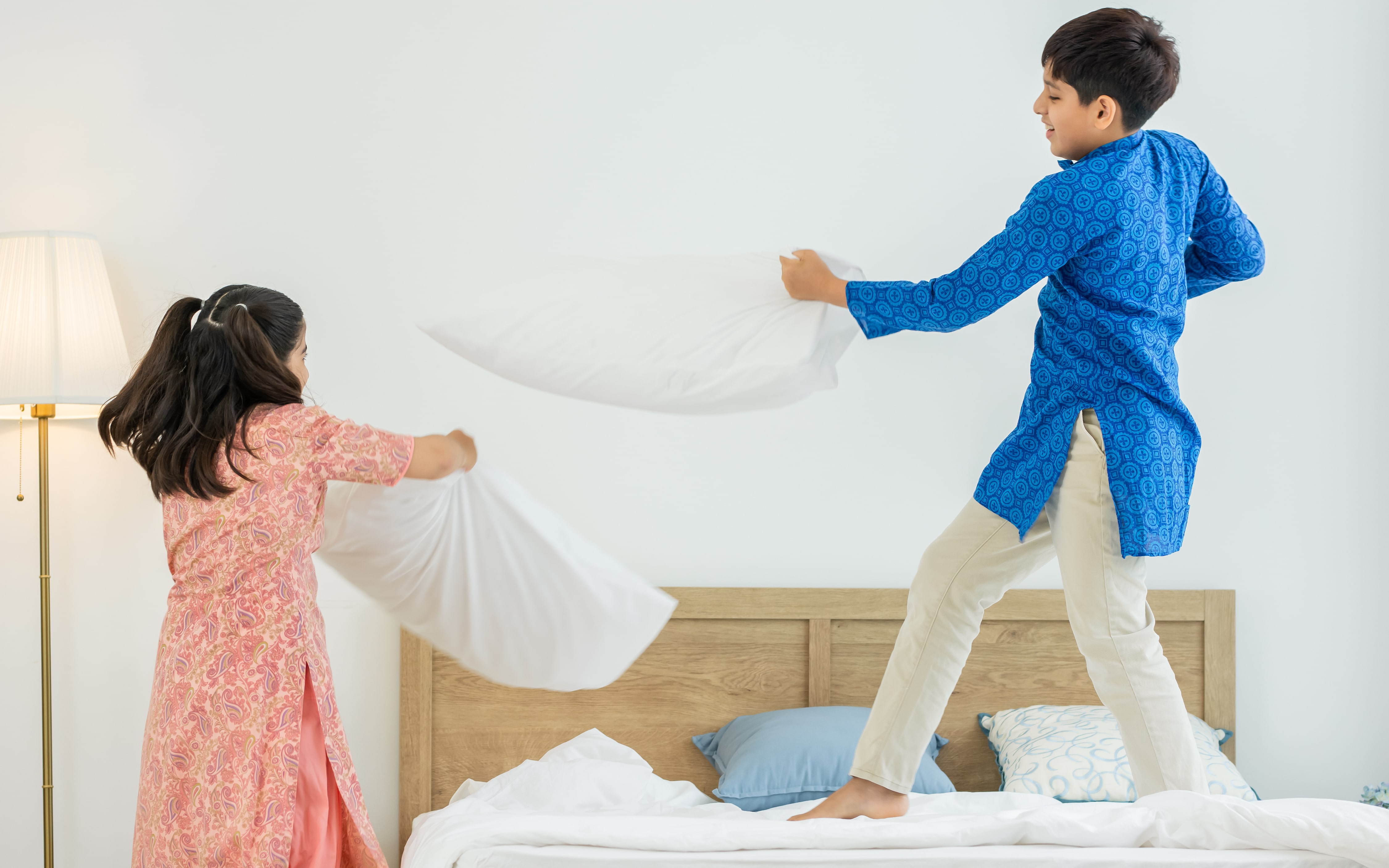 A brother and sister having a pillow fight in a white, blank canvas of a bedroom.