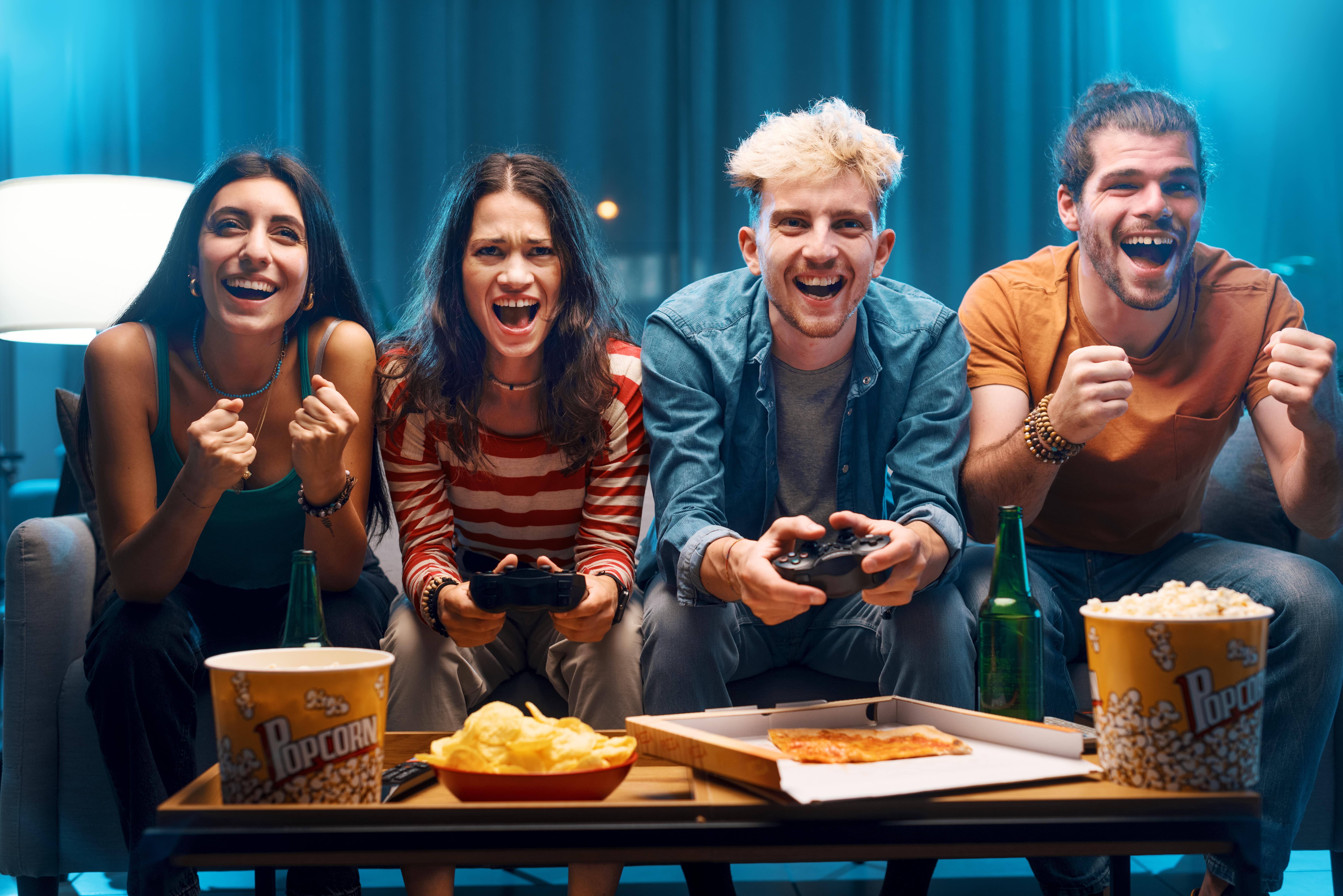 A group of four friends smiling and gaming together with pizza, popcorn, and beverages at the ready.