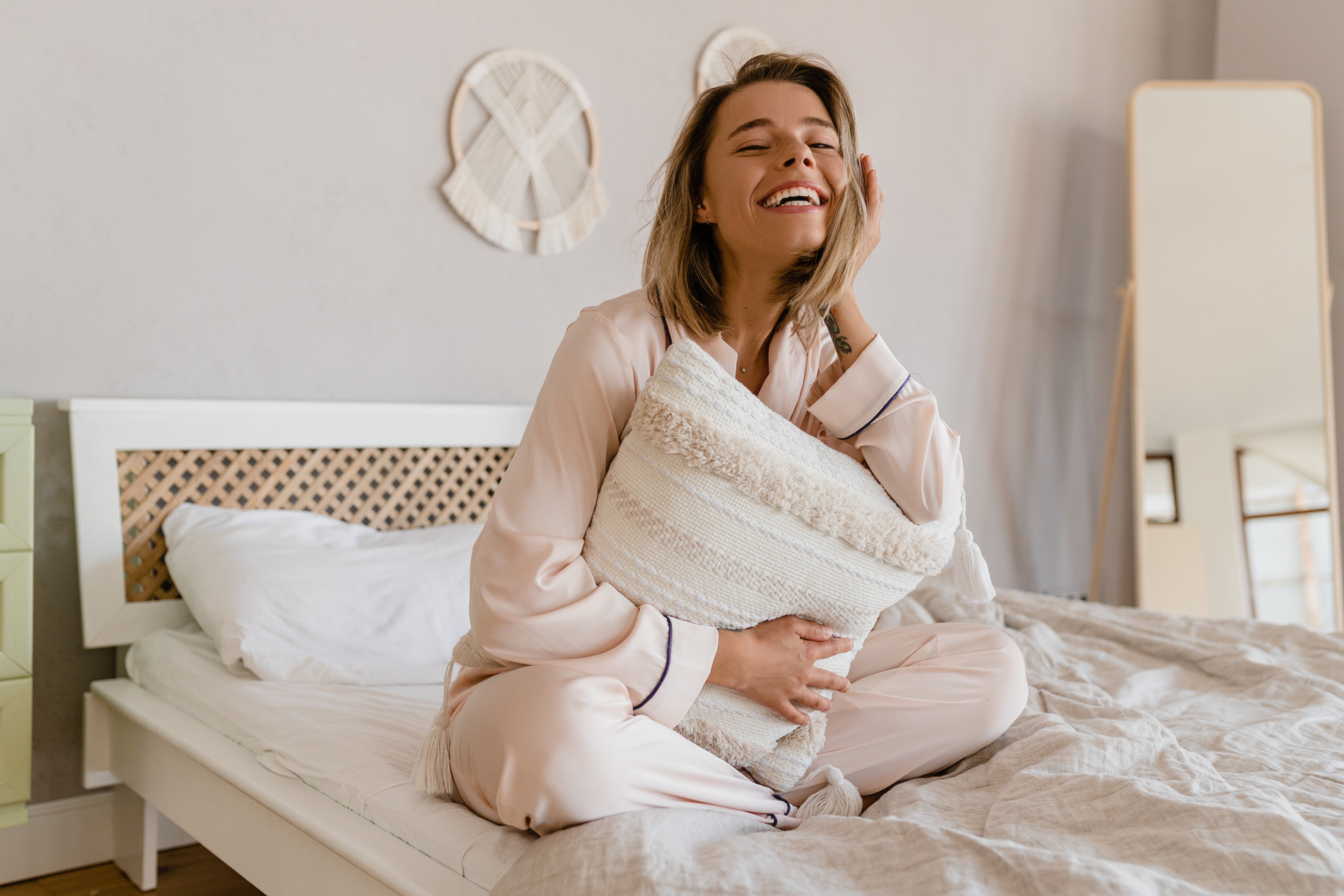 A woman sat crossed legged on the bed smiling and cuddling a throw cushion while wearing new pyjamas.