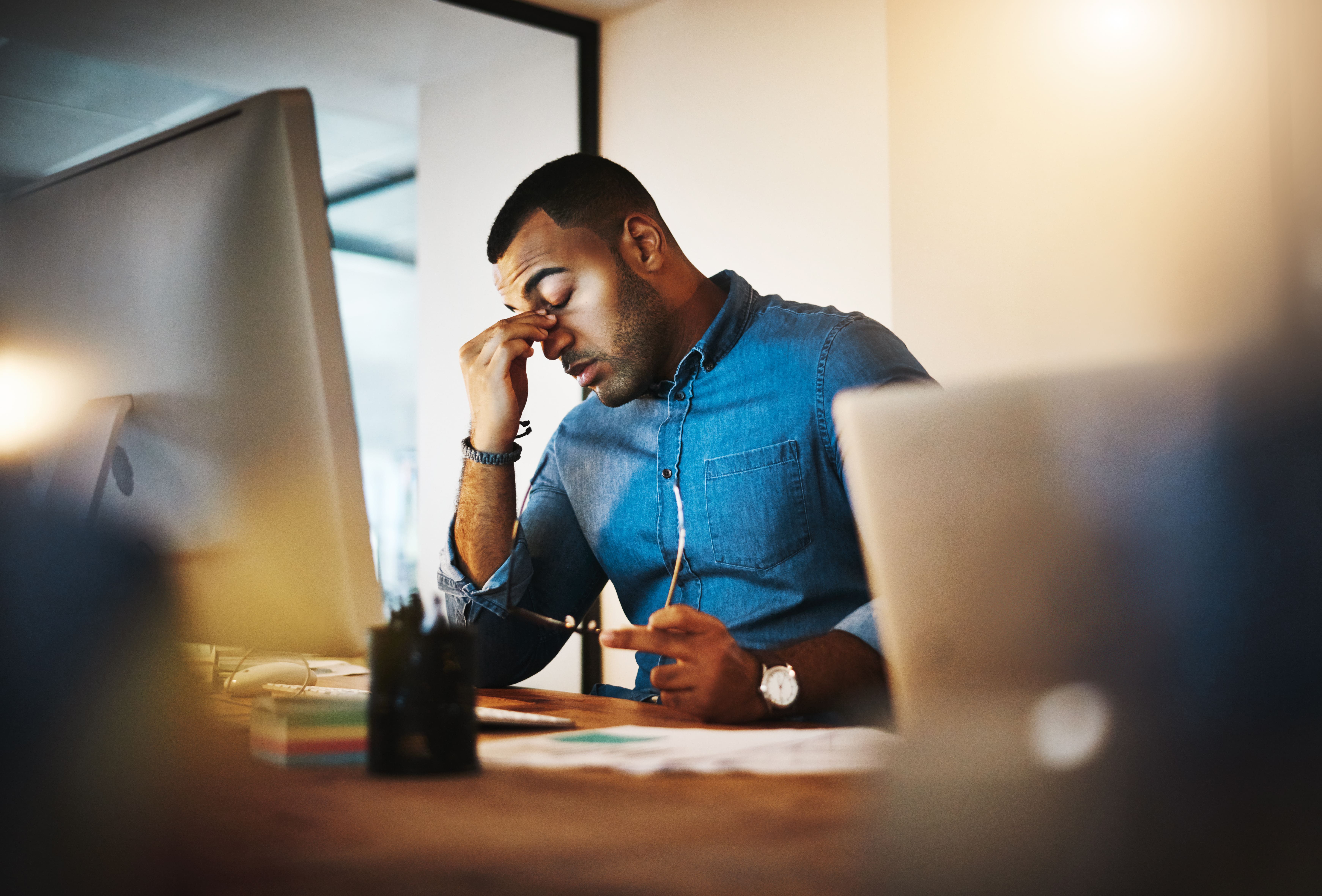 A man sat at his computer rubbing the bridge of his nose as he tries to concentrate while suffering from a lack of sleep.