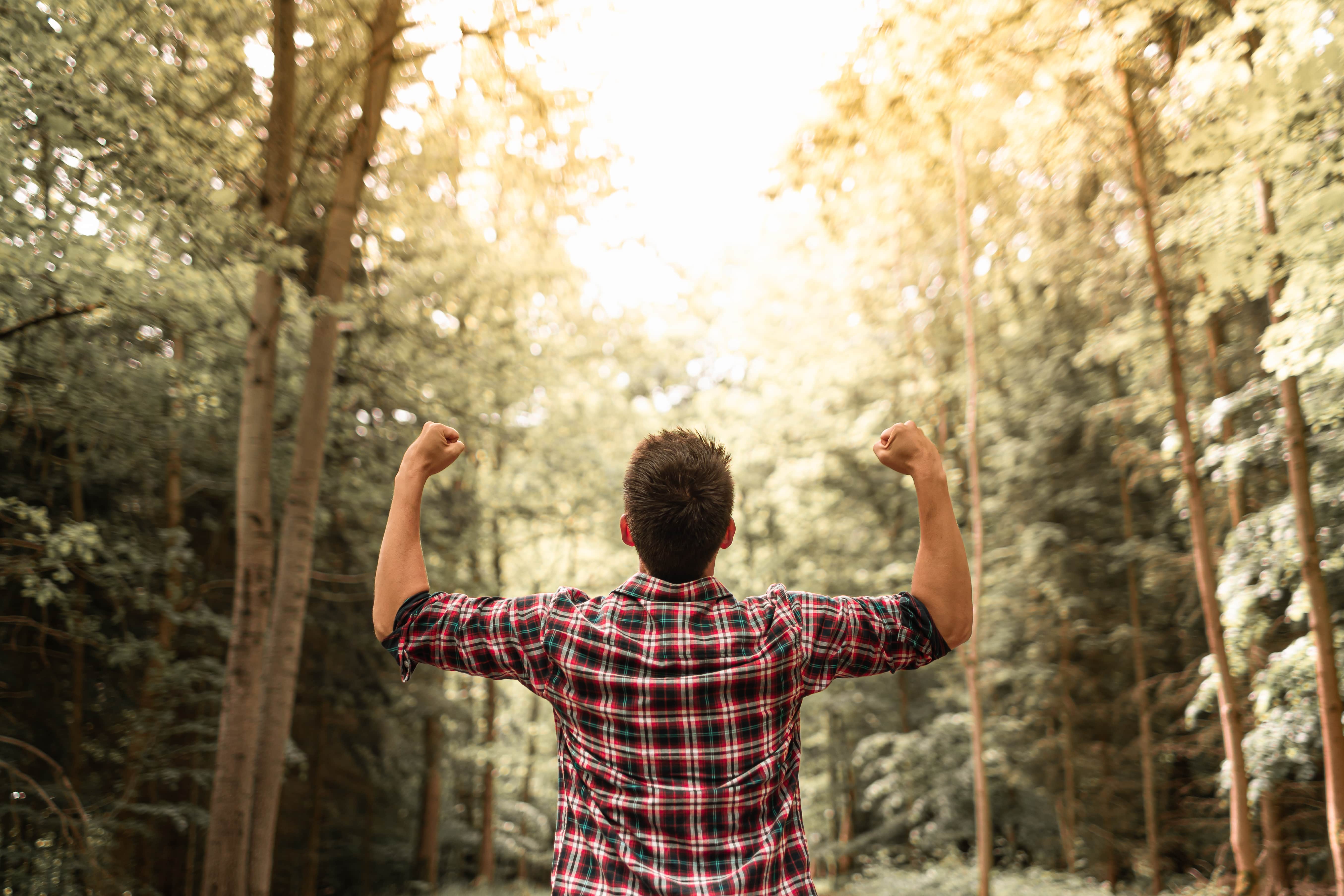 A man stood in the woods with both hand in fists raised towards the sky. This can be read as accomplishment or as a challenge needing to be overcome on the road to achieving his goals.