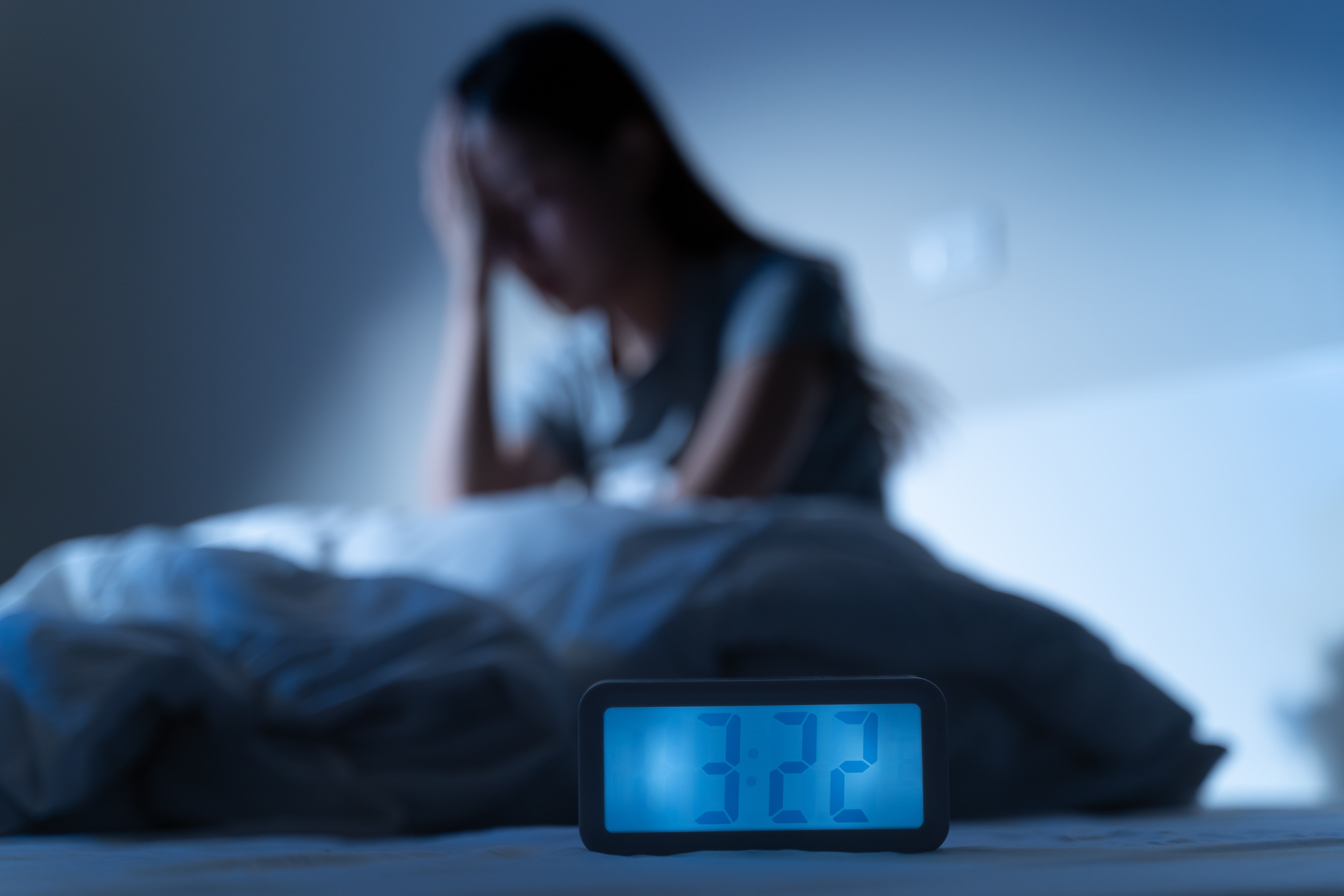 A woman holding her head in her hands as she struggles to get back to sleep after waking up during the night. The time on the digital clock in the foreground of the picture reads 3:22 AM