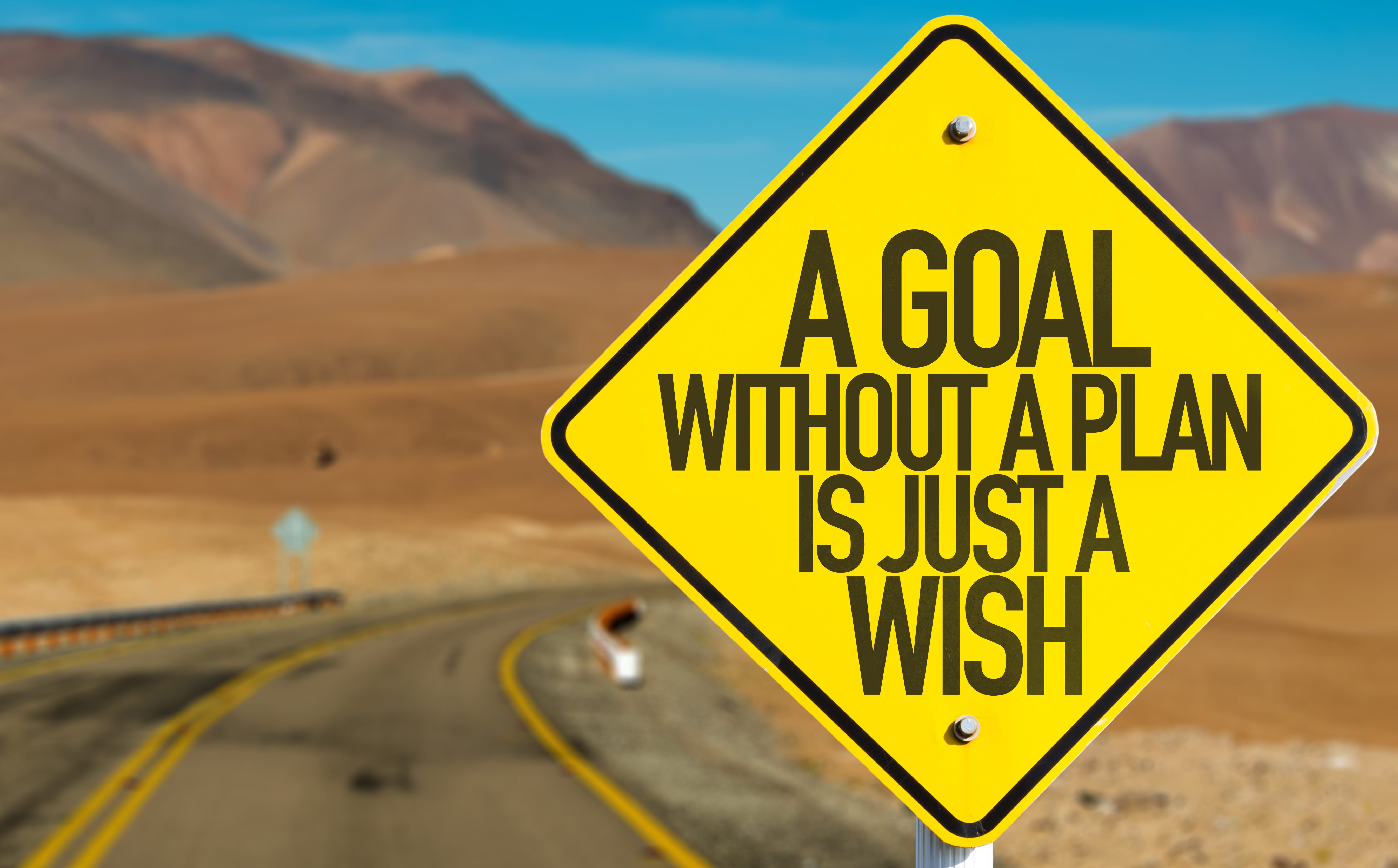 A yellow road sign reading "A goal without a plan is just a wish". In the background, a road leads into a mountainous, desert and the juxtaposes against the bright blue sky.